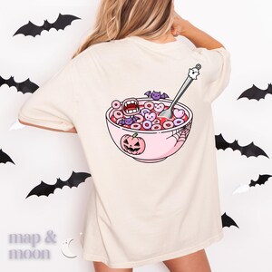May include: Cream-colored t-shirt featuring a Halloween-themed graphic on the back. The design includes a pink bowl of cereal with spooky elements like bats, a pumpkin, and a spiderweb. A spoon with a ghost on top is in the bowl.