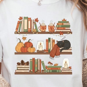 Puede incluir: Camiseta blanca con una ilustración de temática otoñal. El diseño incluye estanterías con libros, calabazas, tazas de chocolate caliente, un gato negro durmiendo y hojas de otoño. La paleta de colores es cálida, con tonos naranja, verde y marrón.