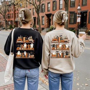 May include: Two sweatshirts, one black and one beige, feature a Halloween-themed design on the back. The design depicts bookshelves with pumpkins, ghosts, black cats, and candles. The women are wearing blue jeans and have their hair up.