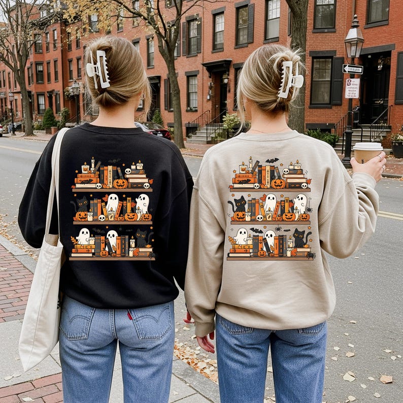 May include: Two sweatshirts, one black and one beige, feature a Halloween-themed design on the back. The design depicts bookshelves with pumpkins, ghosts, black cats, and candles. The women are wearing blue jeans and have their hair up.