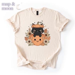 May include: Cream-colored t-shirt featuring a cartoon black cat inside a pumpkin. The pumpkin has a smiling face and is surrounded by fall leaves and flowers. The shirt has the text "map & moon" in the upper left corner.