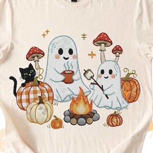 Può includere: T-shirt color crema con un design a tema Halloween. Due fantasmi dei cartoni animati arrostiscono marshmallow su un falò. Un gatto nero è seduto su una zucca. Anche altre zucche e funghi sono nel design.