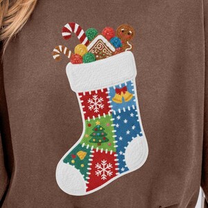 May include: A brown sweatshirt featuring a colorful Christmas stocking design. The stocking is filled with gingerbread cookies, candy canes, and ornaments. The stocking has a white cuff and is decorated with snowflakes, a Christmas tree, and bells.