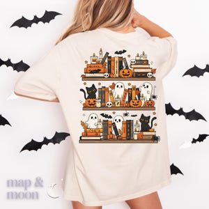 May include: Cream-colored t-shirt featuring a Halloween-themed bookshelf design. The graphic includes stacked books, pumpkins, ghosts, black cats, candles, and bats. The text "map & moon" is at the bottom left.