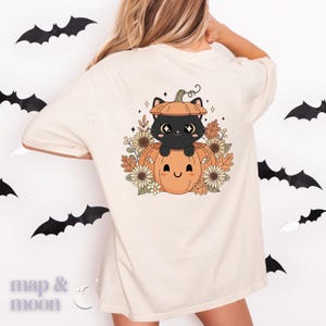 May include: Cream-colored t-shirt featuring a Halloween-themed graphic. The design includes a black cat inside a pumpkin, surrounded by sunflowers and leaves. The shirt has a relaxed fit and is suitable for casual wear.