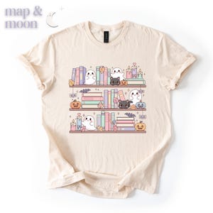 May include: Cream-colored t-shirt featuring a whimsical illustration of a bookshelf with books, ghosts, black cats, pumpkins, candles, and autumn leaves. The text "map & moon" is in the upper left corner. The design is in soft pastel colors.