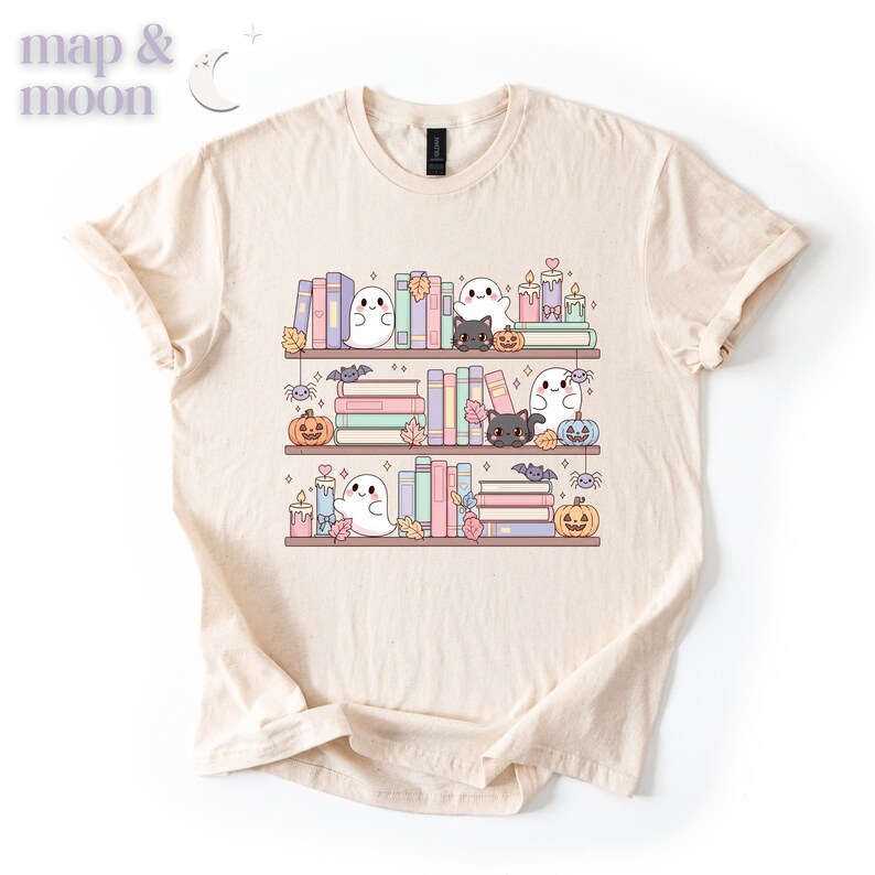 May include: Cream-colored t-shirt featuring a whimsical illustration of a bookshelf with books, ghosts, black cats, pumpkins, candles, and autumn leaves. The text "map & moon" is in the upper left corner. The design is in soft pastel colors.