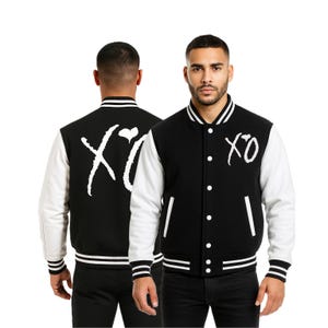 May include: Black and white varsity jacket with white sleeves. The jacket features the text "XO" in white on the front and back. The jacket has a button-up front, striped cuffs, and a striped hemline.