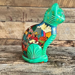 May include: A hand-painted ceramic cat figurine. The cat is primarily green with a detailed, colorful scene painted on its body. The scene depicts figures, buildings, and foliage. The cat is posed in a seated position.
