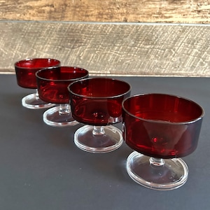 May include: Four ruby red glass dessert bowls with clear stems and bases. The bowls have a cylindrical shape and are arranged in a row. The glass has a glossy finish, reflecting light. The background is a mix of gray and wood.