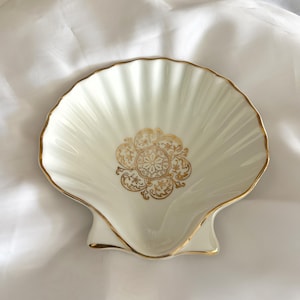 May include: A seashell-shaped trinket dish in cream with gold trim. The dish features a gold floral design in the center. The dish is made of ceramic or porcelain and is likely used for jewelry or small items.