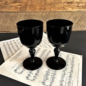May include: Two black wine glasses with a rounded bowl and a short stem. The glasses sit on sheet music with musical notes. The background is a wooden surface.