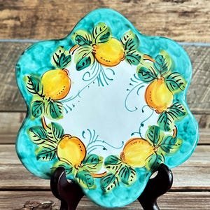 May include: A decorative ceramic plate with a scalloped edge and a turquoise border. The plate features a hand-painted design of yellow lemons and green leaves. The plate is displayed on a dark wooden stand.