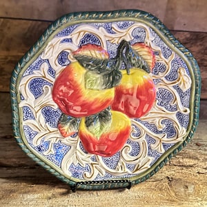 May include: Decorative ceramic plate with a raised relief design of three apples. The apples are red and yellow with green leaves, set against a blue and white background with a gold trim. The plate is octagonal and has a green border.
