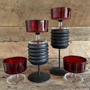 May include: Four decorative candle holders with a unique design. The candle holders feature a clear glass base, a black, accordion-style middle section, and a deep red glass cup. The candle holders are on a wooden surface.