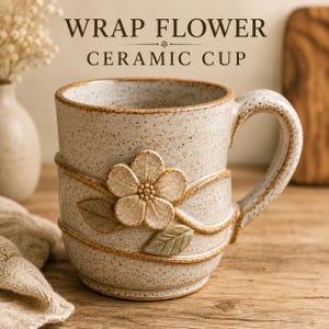 May include: A speckled beige ceramic cup featuring a raised relief design of a five-petaled flower and two leaves wrapped by a thin vine. The text WRAP FLOWER CERAMIC CUP is printed above the cup. The mug has a sturdy handle and a textured finish.