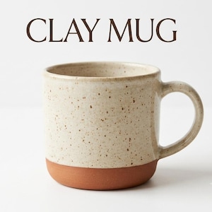 Op de afbeelding: Een keramische mok met de tekst CLAY MUG erboven gedrukt. De mok heeft een gespikkeld crèmekleurig bovengedeelte en een effen terracotta-kleurige basis. Het heeft een cilindrisch lichaam en een enkele gebogen handgreep aan de rechterkant.