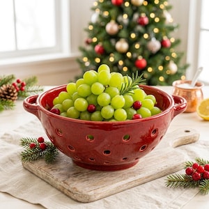 May include: A deep red ceramic colander with two side handles and perforated holes, filled with a large mound of green grapes, a sprig of fresh rosemary, and scattered red cranberries, resting on a wooden cutting board.