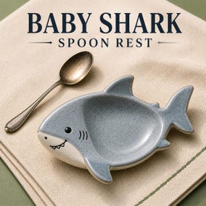 May include: A ceramic spoon rest shaped like a shark with a speckled blue-gray finish, a white underbelly, and a small black eye and mouth. The text BABY SHARK SPOON REST is printed above the shark, accompanied by a metallic bronze-toned spoon.