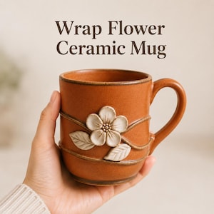 May include: A terracotta-colored ceramic mug featuring the text Wrap Flower Ceramic Mug. The mug has a raised, white, five-petaled flower relief with two leaves and a vine-like design wrapping around the exterior surface of the vessel.