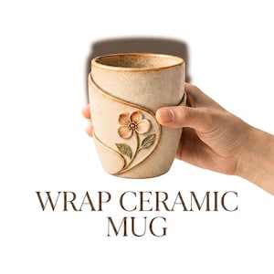 May include: A beige ceramic mug with a wrap-around design and a raised, sculpted flower with pink petals and green leaves on the front. The mug features a speckled texture and a brown rim. The text WRAP CERAMIC MUG is printed below the object.