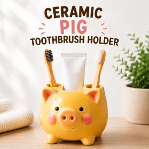 Può includere: Un portaspazzolino in ceramica gialla a forma di maialino con guance rosa, contenente due spazzolini in bambù e un tubetto di dentifricio bianco. Il testo CERAMIC PIG TOOTHBRUSH HOLDER è stampato sopra in lettere marroni e rosa.