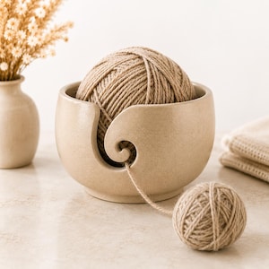 Può includere: Una ciotola per lana in ceramica beige con finitura liscia e maculata, dotata di un'apertura a spirale sul davanti. Un grande gomitolo di lana color marrone chiaro è posto all'interno, con un filo che passa attraverso l'apertura verso un gomitolo.