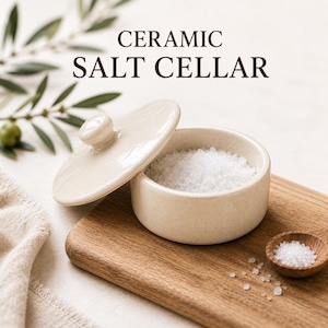 May include: A round, off-white ceramic salt cellar with a matching lid, filled with coarse white salt. The text CERAMIC SALT CELLAR is printed above the container. A small wooden spoon containing salt rests nearby, with scattered salt grains.