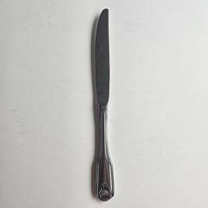Pode incluir: Uma faca de manteiga de aço inoxidável com uma lâmina curva e um cabo decorativo. A alça apresenta um design em forma de concha na base. A faca está posicionada verticalmente contra um fundo branco.