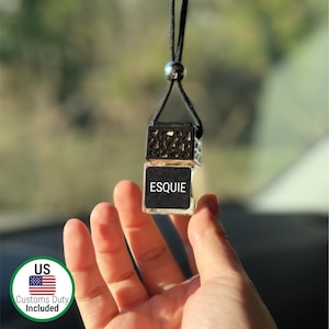 May include: A clear glass car air freshener with the word "ESQUIE" on a black label. The air freshener is suspended by a black cord and metal accents. The image includes a Customs Duty Included sticker.