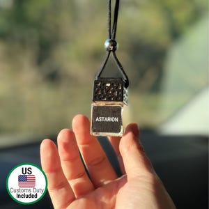 Puede incluir: Un pequeño ambientador para coche de cristal transparente con el nombre "Astarion" impreso en una etiqueta negra. El ambientador está suspendido por un cordón negro y una cuenta plateada. La parte superior es una jaula decorativa de metal negro.