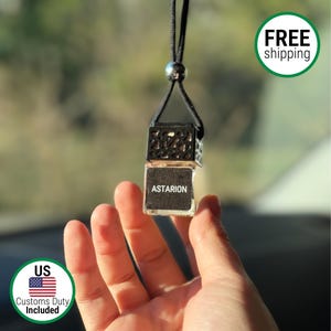 May include: A clear glass car air freshener with the name "Astarion" printed on a black label. The air freshener is suspended by a black cord and a silver bead. The top is a decorative black metal cage.