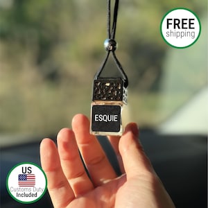 May include: A small, clear glass car air freshener with the word "ESQUIE" printed on a black label. The air freshener is suspended by a black cord and metal bead. The product is held in a hand.