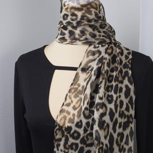 Può includere: Una sciarpa trasparente con stampa leopardata drappeggiata su un top nero a maniche lunghe. La sciarpa presenta un motivo di macchie marroni e nere su sfondo beige chiaro. Il top ha uno scollo a chiave.