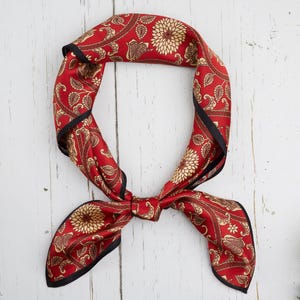 May include: Red silk scarf with a floral and paisley pattern. The scarf has a black border and is tied in a knot. The scarf is approximately 69 cm long.