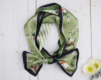 Könnte beinhalten: Ein Seidenschal in Grün-, Schwarz- und Weißtönen, verziert mit einem Blumenmuster. Der Schal hat eine Schleife und einen kontrastierenden schwarzen Rand. Auf weißer Holzoberfläche.