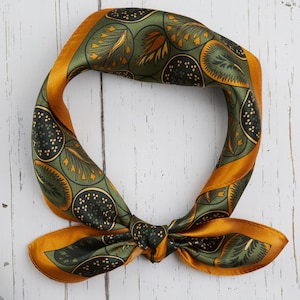 Può includere: Una sciarpa quadrata di seta con un motivo botanico verde oliva, oro e nero. La sciarpa è annodata, con le estremità aperte. È appoggiata su una superficie di legno bianco.