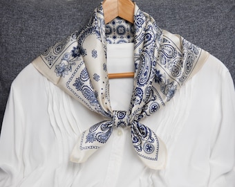 May include: A cream-colored silk scarf with a navy blue paisley pattern is draped over a white blouse. The scarf is knotted at the neck and hangs on a wooden hanger. The blouse has pleats down the front.