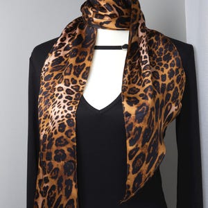 May include: A leopard print scarf draped over a black long-sleeve top. The scarf features a repeating pattern of brown and black spots on a tan background. The top has a V-neck and a black choker.