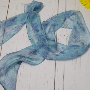 May include: A light blue scarf with a feather pattern in shades of blue and purple. The scarf is made of a sheer fabric and is draped on a white wooden surface. A yellow flower is in the upper right corner.