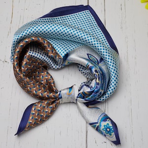 May include: A square silk scarf with a variety of patterns and colors. The scarf features geometric shapes in brown and blue, a blue and white grid pattern, and a floral design. The scarf is tied in a knot.