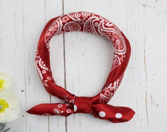 May include: A red bandana with a white paisley pattern tied in a knot. The bandana is made of a silky material and has white polka dots on the ends. The background is a white wooden surface.