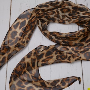 Può includere: Una sciarpa trasparente con stampa leopardata nei toni del marrone, beige e nero. La sciarpa è drappeggiata su una superficie di legno bianco. Un fiore giallo è nell'angolo in basso a destra.