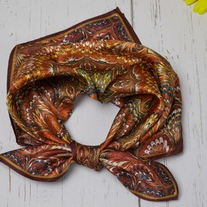 May include: A patterned silk scarf in shades of brown, orange, and yellow, with a dark burgundy border. The scarf is knotted and made of a soft, flowing material. The abstract design features intricate details.