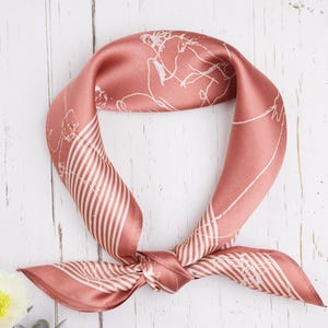 May include: A dusty rose silk scarf with white linear and abstract designs. The scarf is knotted at the bottom and has a striped pattern on the sides. The scarf is on a white wooden surface.