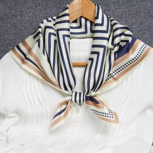 May include: A white blouse with pleats is adorned with a silk scarf. The scarf features a geometric pattern in navy blue, cream, and tan. The scarf is knotted at the neck, creating a stylish accessory.
