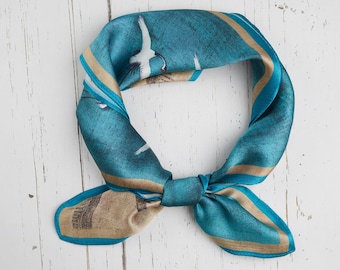 May include: A teal and tan silk scarf with a nautical design. The scarf features a blue background with white birds and tan accents. The scarf is tied in a knot, showcasing its design and texture. The scarf is made of a soft, silky material.
