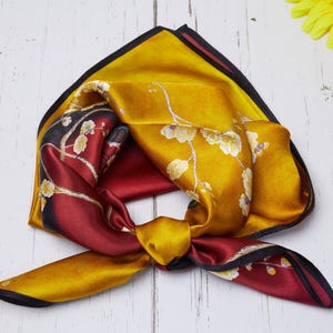 Puede incluir: Un pañuelo cuadrado de seda con un estampado floral. El pañuelo presenta una paleta de colores dorados, burdeos y negros. El pañuelo está anudado en el centro, mostrando los diferentes patrones y colores.