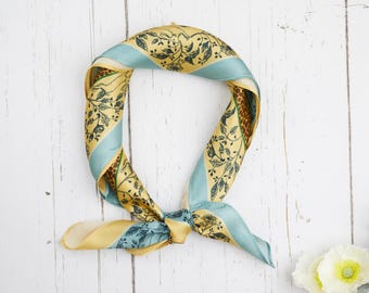 May include: A silk scarf with a botanical pattern in shades of yellow, teal, and brown. The scarf is tied in a knot and lies on a white wooden surface. The scarf's design features leaves and vines.