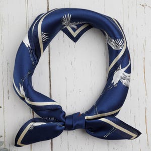 May include: A navy blue silk scarf with a white crane pattern. The scarf is knotted and has a square shape. The background is a white wooden surface.